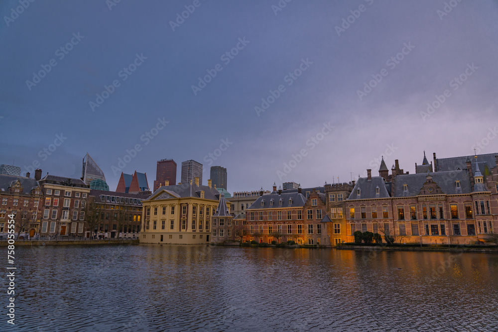 Fototapeta premium The Hague, Netherlands March 22 2023: Binnenhof castle (Dutch Parliament) with the Hofvijver lake against a background of skyscrapers and blue cloudy sky at dusk (Den Haag)