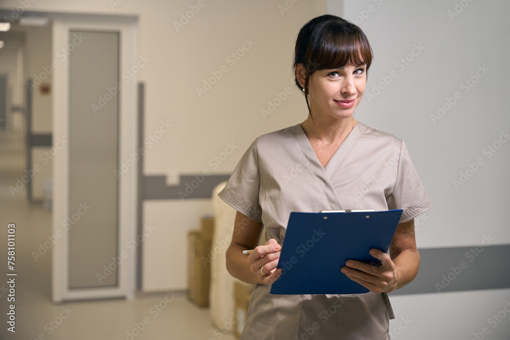 Female medical worker holding clipboard standing in hospital hallway Female medical worker holding clipboard standing in hospital hallway
