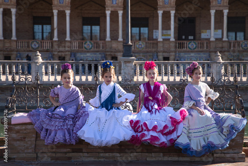 Four little girls dancing flamenco dressed in typical flamenco dress sitting on a bench in a famous square in seville, spain. In the background the arches and columns of the famous square.
