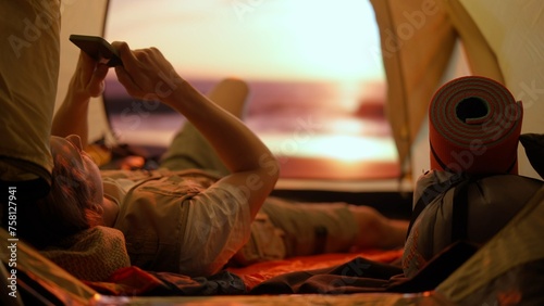 Person on campsite traveling and hiking, exploring nature. Young man laying texting on smartphone inside the tent at sunset on the beach, holiday and vacation outdoors
