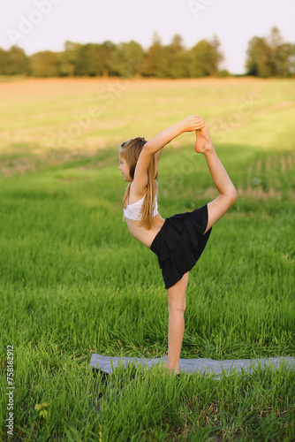 Little cute girl in white top and black skirt-shorts practicing yoga pose on amat on nature backround at summer. Healthy lifestyle concept photo