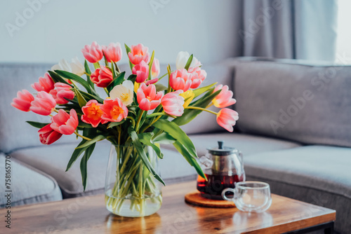 Focus on vase of fresh tulips bouquet and just brewed tea pot and cup on coffee table with gray couch sofa in modern leaving room. Cozy, calm and comfortable relax at home. Time to take a break. #758165770