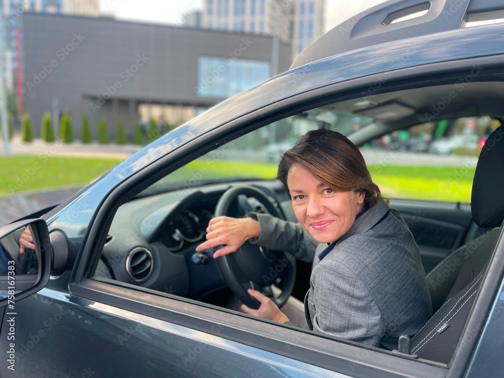 Close-up of beautiful cheerful businesswoman sitting in own car looking at camera and smiling. Positive Caucasian female driving a vehicle. Urban lifestyle. Business concept