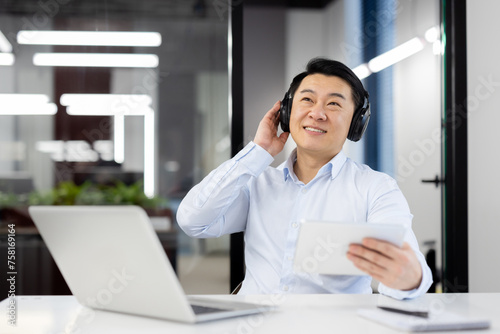 Relaxed young Asian man sitting in the office at the desk wearing headphones, holding a mobile phone, listening to music. rests during a work break.