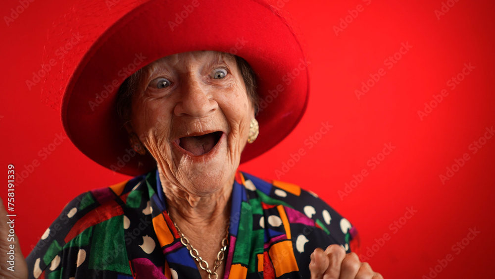 Funny fisheye of elderly senior old woman with wrinkled skin wearing red hat gives great idea gesture showing explosion of thinking posing isolated on red background.