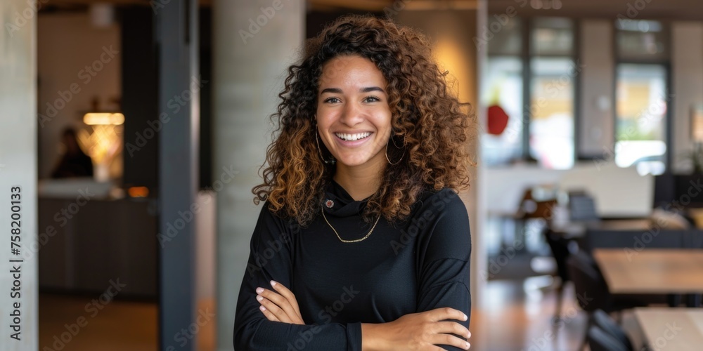 a cheerful young woman with curly hair, smiling broadly at the camera. She stands with her arms crossed and looks confident and friendly. The setting appears to be a casual office space a cheerful young woman with curly hair, smiling broadly at the camera. She stands with her arms crossed and looks confident and friendly. The setting appears to be a casual office space