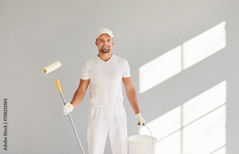 Portrait of a happy smiling male painter in white clothes holding a bucket and paint roller standing in empty room on grey wall background. Renovation, apartment repair and renovating house concept. Portrait of a happy smiling male painter in white clothes holding a bucket and paint roller standing in empty room on grey wall background. Renovation, apartment repair and renovating house concept.