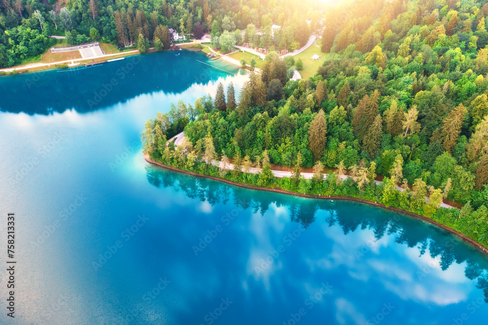 Aerial view of road near blue lake, green forest at sunrise in summer. Bled lake, Slovenia. Travel. Top view of beautiful road, trees in spring. Landscape with highway and sea bay. Road trip. Nature