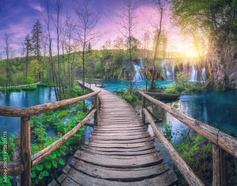 Waterfall and wooden path in green forest. Plitvice Lakes, Croatia at sunset in spring. Colorful landscape with pathway in blooming park, trees, water lilies, river, pink sky in summer. Trail in woods