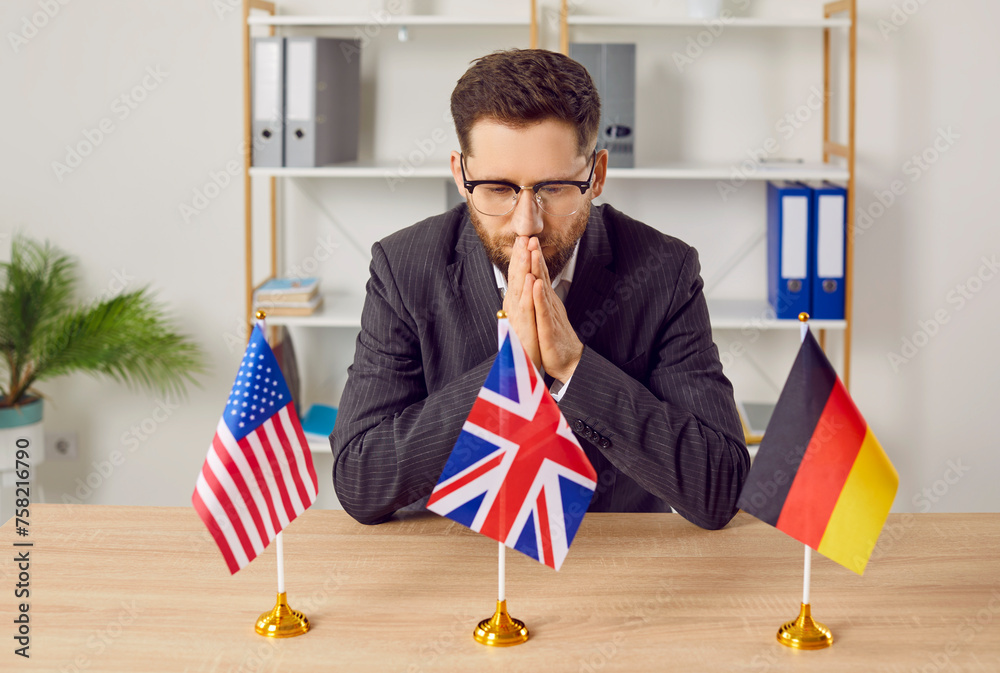 Portrait of a thoughtful pensive young business man in suit sitting at the desk in office with USA, Great Britain and Germany flags choosing country to immigrate or foreign language to study. - obrazy, fototapety, plakaty 