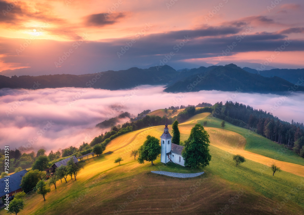 Aerial view of small church on the hill over pink low clouds at foggy sunrise in summer in Slovenia. Top view of beautiful chapel on mountain in fog, green meadows, trees, orange sky at dawn in spring