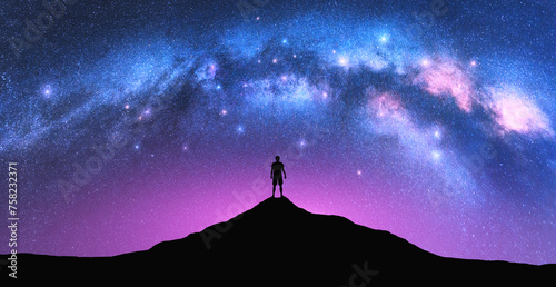 Milky Way arch and man on the mountain peak at starry night. Silhouette of alone guy, pink sky with bright stars in summer. Galaxy. Space background. Landscape with arched milky way. Travel and nature