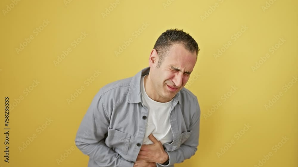 Hispanic young man wearing shirt clutches stomach in painful ache, ill with stomach illness. stands isolated on yellow background displaying ache in abdominal area. Hispanic young man wearing shirt clutches stomach in painful ache, ill with stomach illness. stands isolated on yellow background displaying ache in abdominal area.