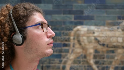 Berlin, Germany, August 16, 2023. Portrait of a young caucasian man with curly hair and glasses busy listening to the audio guide with headphones at the Ishtar Gate in the Pergamon Museum in Berlin. photo