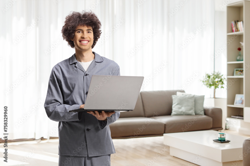 Young man in pajamas standing in a living room with a laptop computer Young man in pajamas standing in a living room with a laptop computer