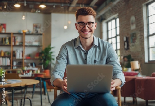 Cheerful young man with glasses using laptop in a cafe. Casual work environment with people in the background.