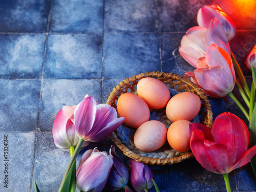 Easter colored eggs in a basket, a bouquet of pink and purple tulips on a blue table. Copy space happy Easter poster
