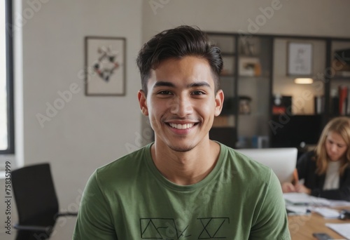 A cheerful young man in a green shirt smiles in a casual office setting. Colleagues work in the background.
