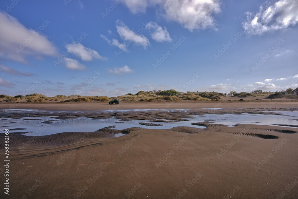 Majestic image of a beach with a tractor driving in the distance, blue sky with white clouds, puddles of water on the sand and dunes Majestic image of a beach with a tractor driving in the distance, blue sky with white clouds, puddles of water on the sand and dunes