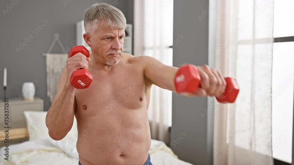 Mature gray-haired man exercises with dumbbells in a bedroom, showing determination and fitness at home. Mature gray-haired man exercises with dumbbells in a bedroom, showing determination and fitness at home.