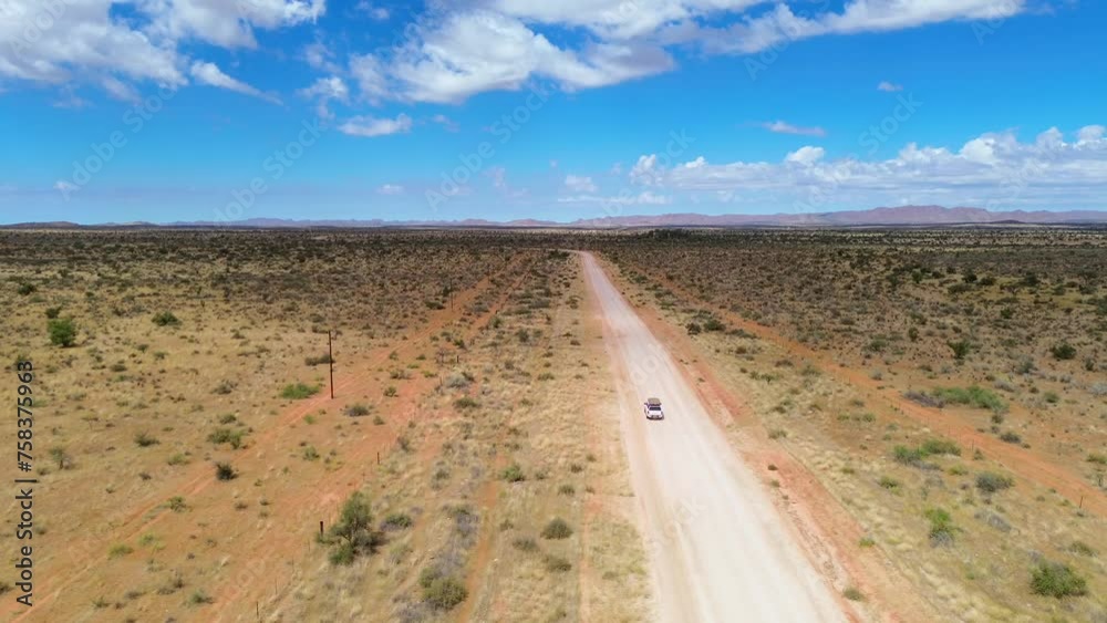 Aerial drone film of a single white safari car, offroad vehicle driving on a narrow dirt or asphalt gravel road through desert landscape. Red earth and high sand dunes near Sossusvlei, Namibia, Africa
