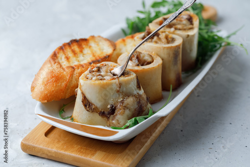 Homemade Roasted Beef Bone Marrow with spoon and croutons on plate, wooden board grey background. Side View.
