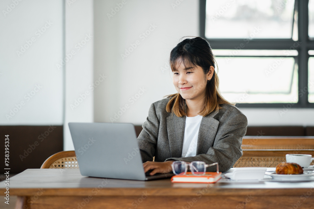 Smiling young businesswoman working on her laptop at a desk, with glasses and a croissant on the table in an airy office. Smiling young businesswoman working on her laptop at a desk, with glasses and a croissant on the table in an airy office.
