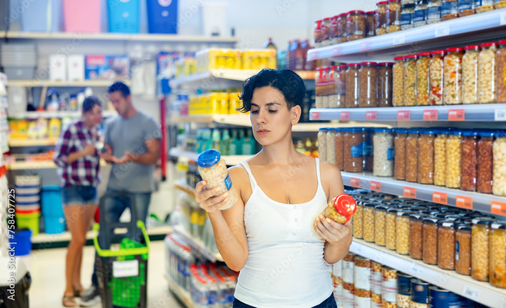 Latin woman standing at shelf in grocery store and selecting pickled products.