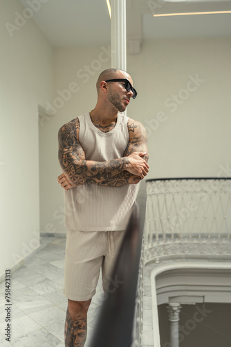 Chico joven musculoso y tatuado posando en hall de hotel moderno con ropa urbana estilosa