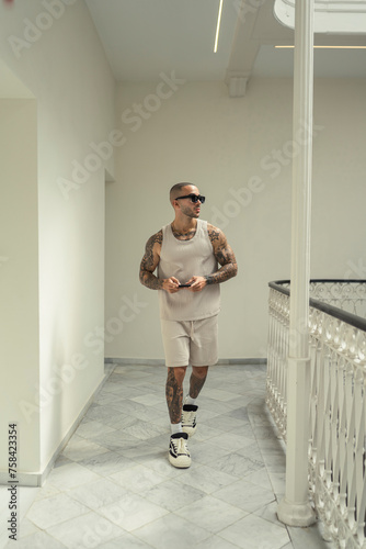 Chico joven musculoso y tatuado posando en hall de hotel moderno con ropa urbana estilosa