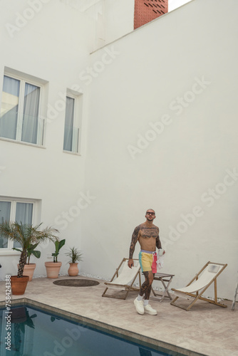 Chico joven tatuado y fitness posando en bañador en piscina de hotel 