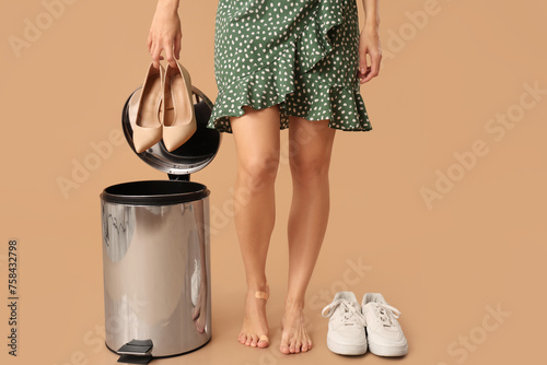 Young woman throwing high-heeled shoes into trash bin on beige background. Uncomfortable shoes concept
