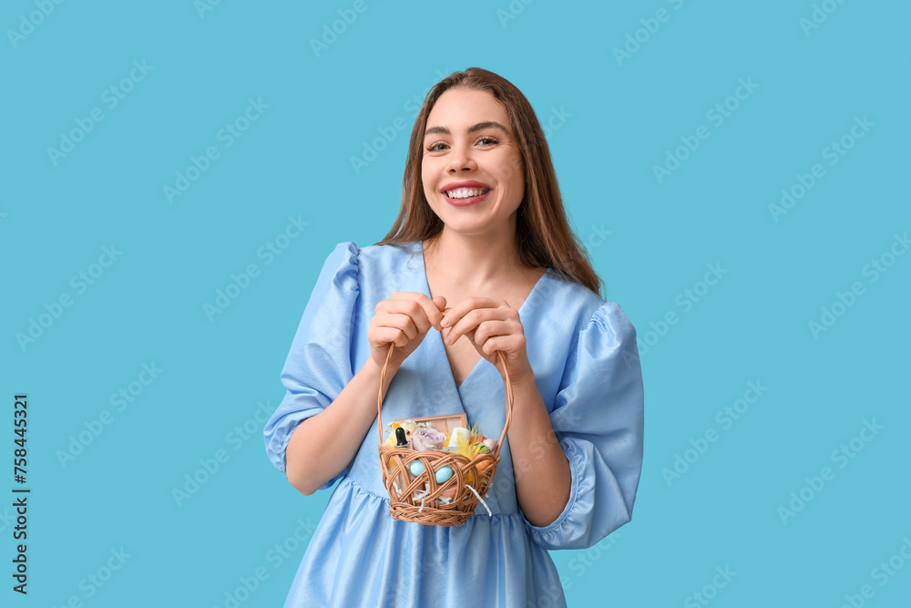 Happy smiling young woman holding basket with makeup products, flowers and Easter decor on blue background Happy smiling young woman holding basket with makeup products, flowers and Easter decor on blue background
