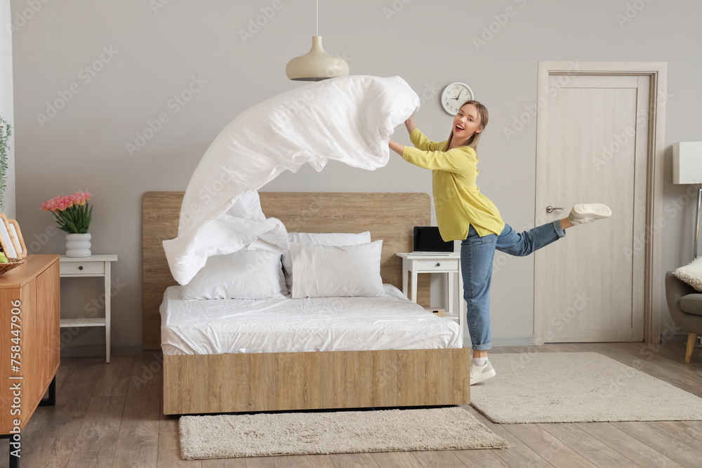 Cheerful young woman making bed in modern bedroom - obrazy, fototapety, plakaty 