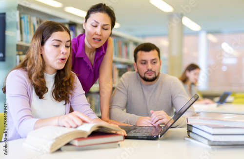 Three students are preparing for classes on a laptop in the university library, where girls classmates are looking for ..information in a textbook and discussing something