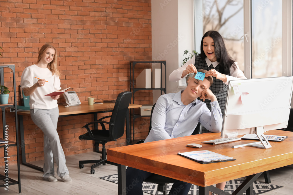 Young woman sticking paper on her sleeping colleague's forehead in office. April Fools' Day celebration Young woman sticking paper on her sleeping colleague's forehead in office. April Fools' Day celebration