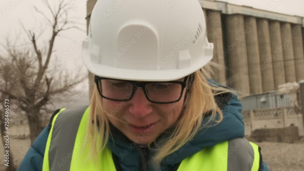 Сlose-up of smiling female contractor in glasses and hard hat in a protective helmet looking at documents in wind against backdrop of an industrial production factory in remote area.