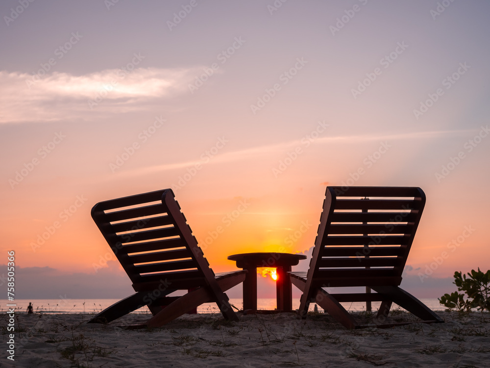 Two empty silhouette wooden sunbeds, couple chair seat and side table on sand beach, peaceful evening sunset sky background with copy space, holiday or summer vacation concept.