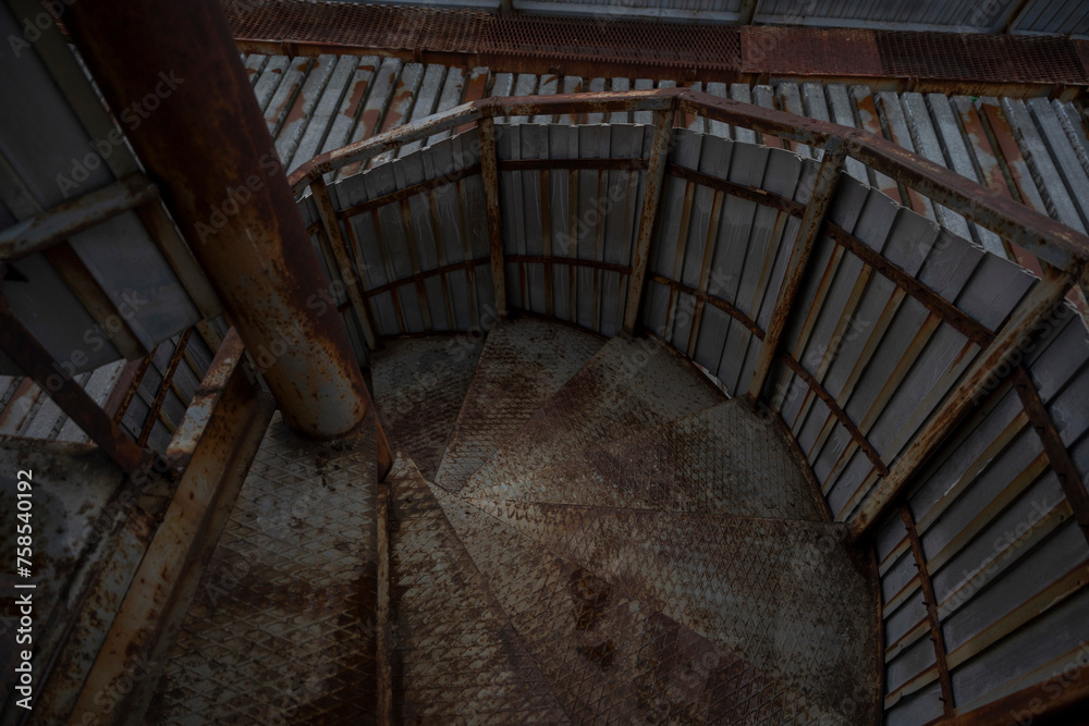Obraz premium Weathered metal stairs spiral upward inside an industrial structure, hinting at the passage of workers past. The wear and rust speak to the harsh conditions and the relentless passage of time.
