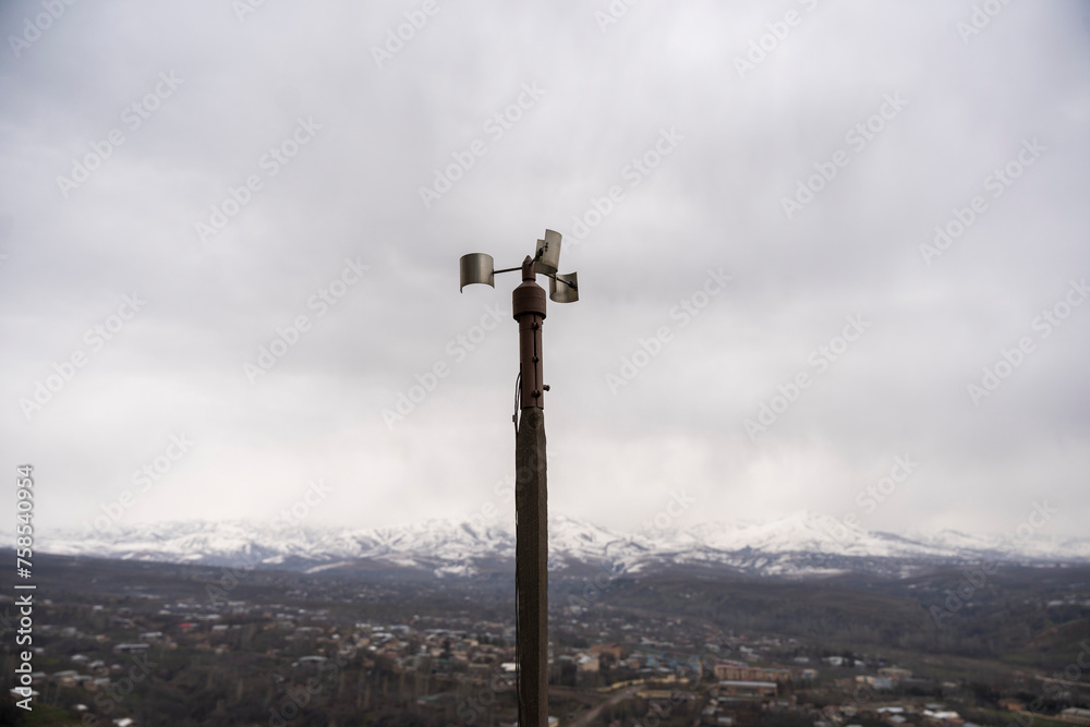 A solitary wind sensor perched atop a slender pole, stark against an overcast sky, silently monitors atmospheric conditions above an industrial complex. Its simplicity belies its importance in data co