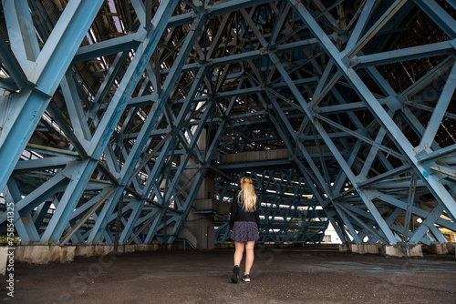 Tableau sur toile A lone figure stands underneath the imposing blue steel lattice of an industrial structure, the geometric patterns creating a cathedral-like vault above