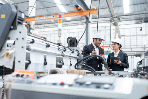 An American male engineer and an Asian female executive inspect the production of materials from machines in an automobile factory. Wear a safety helmet and suit. Use a laptop for work