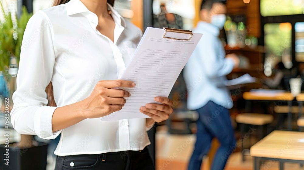 Closeup of a person holding documents with a clipboard in a blurred cafe setting suggesting a business or service environment