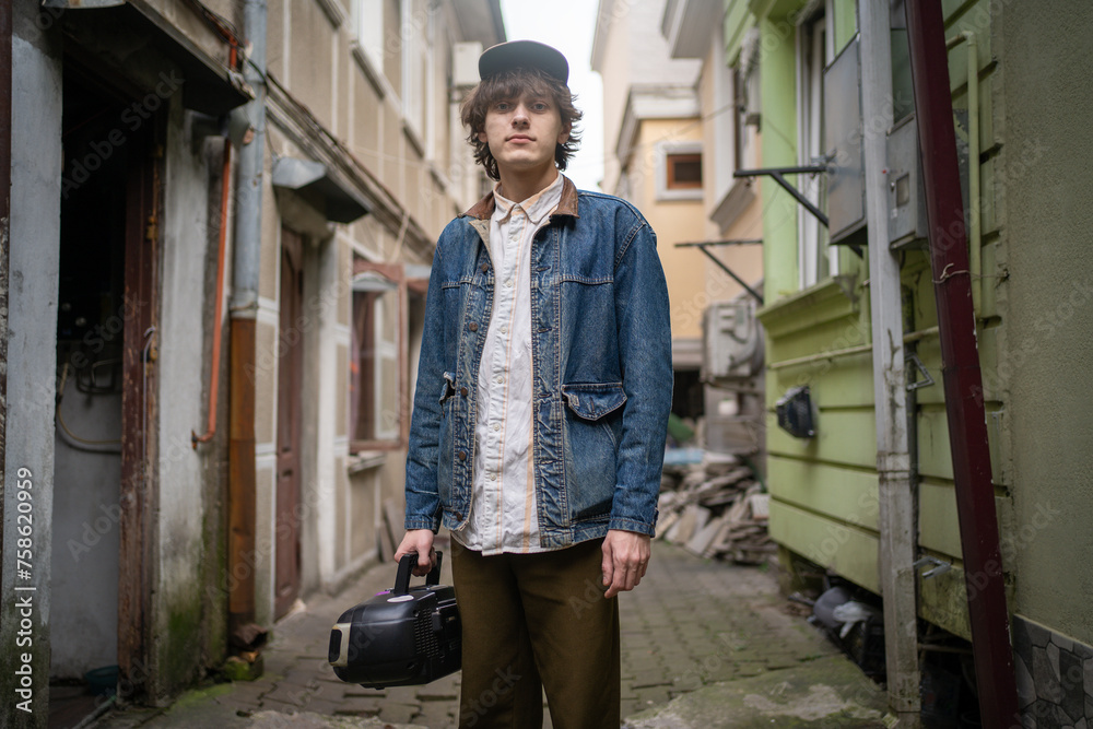 Fashionable young man in 90s wear style holding vintage tape cassette player, walks through narrow alley in an old town setting. Fashionable young man in 90s wear style holding vintage tape cassette player, walks through narrow alley in an old town setting.
