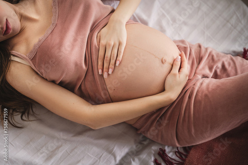 a pregnant woman in pajamas lies on the bed and strokes her big belly. Waiting for the baby. Close-up of a hand without a face