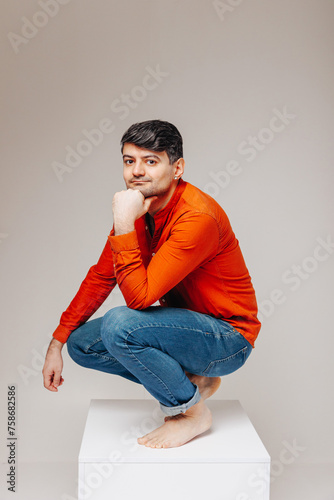 A man squats thoughtfully on a white cube and looks at the camera smiling, the concept of coming up with something