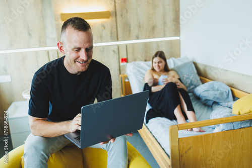 A young man works on a laptop against the background of his wife who is breastfeeding her newborn child. Work online on a laptop.