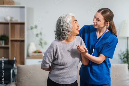Caucasian female doctor assists in supporting an elderly Asian patient while they both sit on the sofa, ensuring comfort and stability during the interaction.