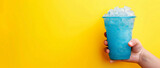 The hand holds a cup with a milk slushie. A  blue glass with a  coconut slushie  on a yellow background, place for text