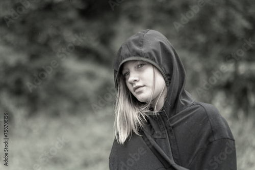 Porträt junges hübsches Mädchen in Regenjacke mit Kapuze. Bei grauem wolkenverhangenen Himmel geht das Mädchen durch den Wald spazieren. Sie hat lange blonde Haare, misstrauisch blickende Augen.